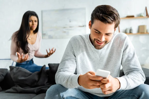 Foco seletivo de homem sorridente usando smartphone e mulher americana africana irritada olhando para ele — Fotografia de Stock