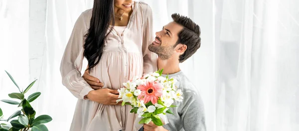 Plan panoramique de bel homme embrassant le ventre de sa femme afro-américaine enceinte et tenant un bouquet — Photo de stock