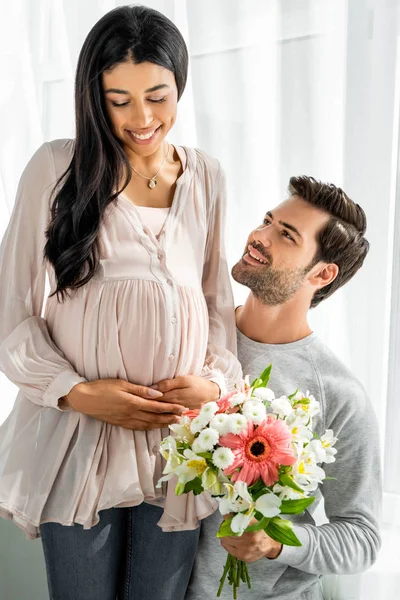 Guapo hombre abrazando vientre de su embarazada africana americana mujer y celebración ramo - foto de stock