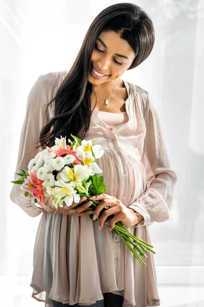 Souriant femme afro-américaine enceinte tenant bouquet dans l'appartement — Photo de stock