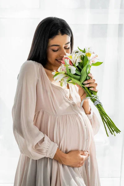 Sonriente embarazada afroamericana mujer oliendo ramo en apartamento - foto de stock