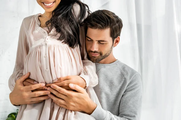 Vista recortada de un hombre guapo abrazando el vientre de su mujer afroamericana embarazada - foto de stock