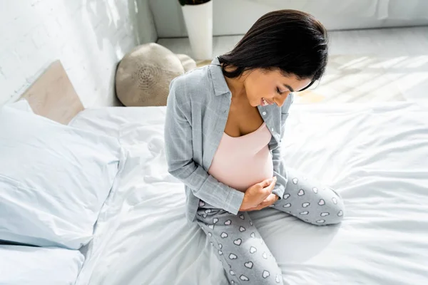 High angle view of african american pregnant woman in pajamas smiling and hugging belly — Stock Photo