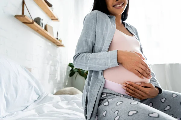 Vue recadrée de la femme enceinte afro-américaine en pyjama souriant et étreignant le ventre — Photo de stock