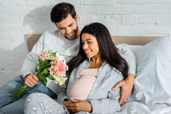 Bel homme tenant bouquet et étreignant sa femme africaine américaine enceinte souriante — Photo de stock