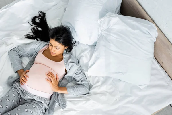 Top view of african american pregnant woman in pajamas touching belly and feeling pain — Stock Photo