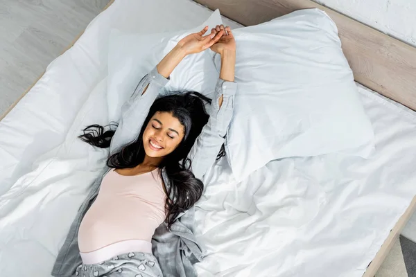 Вид сверху на африканскую беременную женщину в пижаме, улыбающуюся и лежащую в постели — стоковое фото