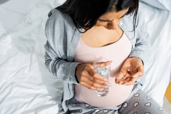 Vista ad alto angolo di donna africana americana incinta in pigiama con pillola e bicchiere d'acqua — Foto stock