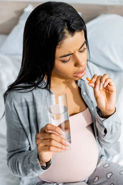 Mulher grávida afro-americana em pijama tomando pílula e segurando vidro de água — Fotografia de Stock