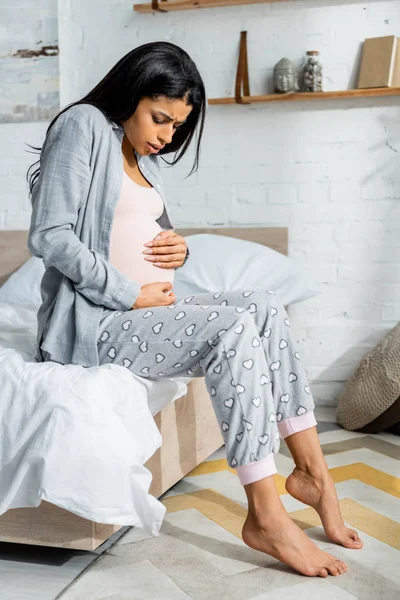 Mujer embarazada afroamericana en pijama abrazando el vientre y sintiendo dolor - foto de stock