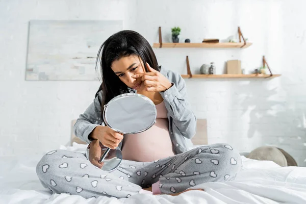 Mujer embarazada afroamericana en pijama mirando el espejo y tocando la cara en el apartamento - foto de stock
