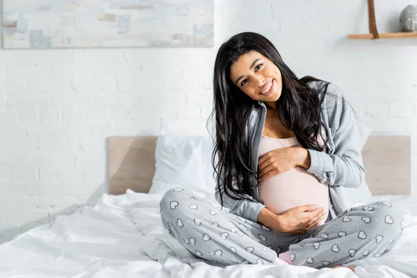Sonriente africano americano embarazada mujer en pijama mirando cámara y abrazando vientre - foto de stock
