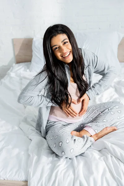 Femme enceinte afro-américaine souriante en pyjama regardant la caméra et montrant un signe cardiaque — Photo de stock