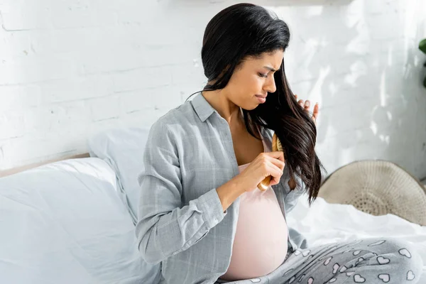 Mujer embarazada afroamericana en pijama cepillándose el pelo en apartamento - foto de stock