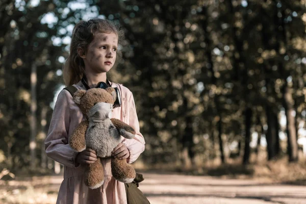 Cute kid holding dirty teddy bear near trees in chernobyl, post apocalyptic concept — Stock Photo
