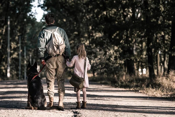 Вид сзади человека, держащего за руки ребенка рядом с немецкой пастушьей собакой, пост-апокалиптическая концепция — стоковое фото