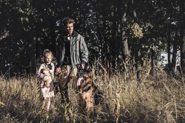 Вродливий чоловік, що стоїть на полі з дитиною та німецьким собакою - пастухом, постапокаліптична концепція. — стокове фото