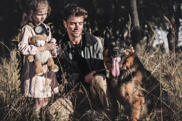 Schöner Mann berührt deutschen Schäferhund in der Nähe von Kind mit Teddybär, postapokalyptisches Konzept — Stockfoto