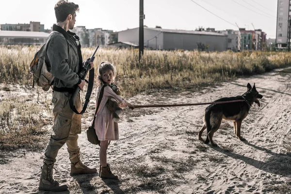 Вооруженный мужчина держит пистолет рядом с ребенком с плюшевым мишкой и собакой, пост-апокалиптическая концепция — стоковое фото