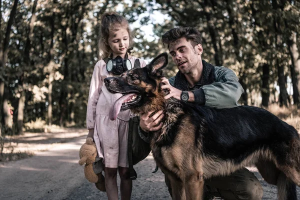 Красивый мужчина трогает немецкую овчарку рядом с ребенком, пост-апокалиптическая концепция — стоковое фото