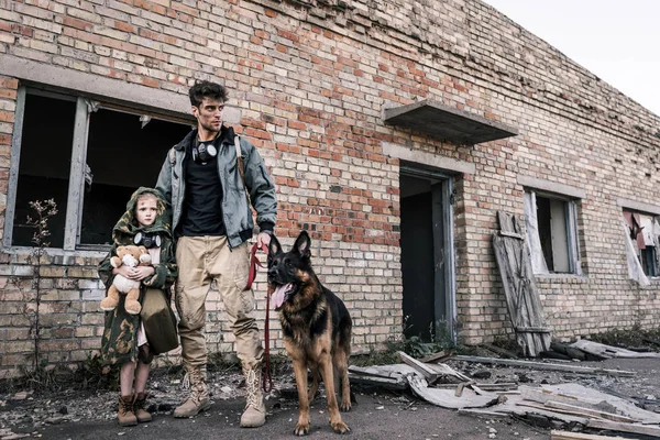 Hombre guapo de pie con niño y perro pastor alemán cerca de edificio abandonado, concepto post apocalíptico - foto de stock