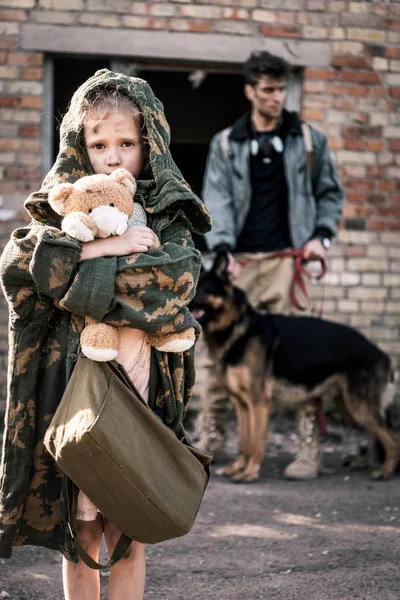 Selektiver Fokus des Kindes mit Teddybär in der Nähe eines Mannes mit Schäferhund in der Nähe eines verlassenen Gebäudes, postapokalyptisches Konzept — Stockfoto