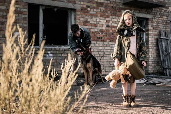 Selective focus of child holding teddy bear near man with german shepherd dog near abandoned building, post apocalyptic concept — Stock Photo