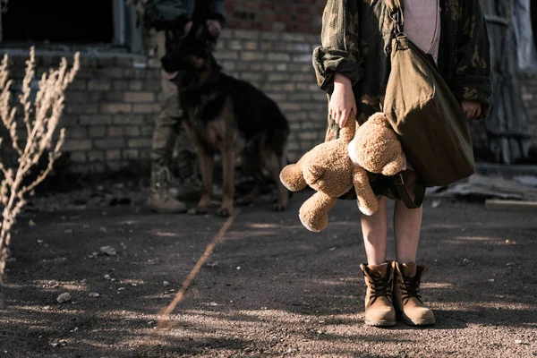 Перекручений вид на дитину, яка тримає Тедді ведмедя поруч з людиною з німецьким пастухом-собакою біля покинутого будинку, постапокаліптична концепція — стокове фото