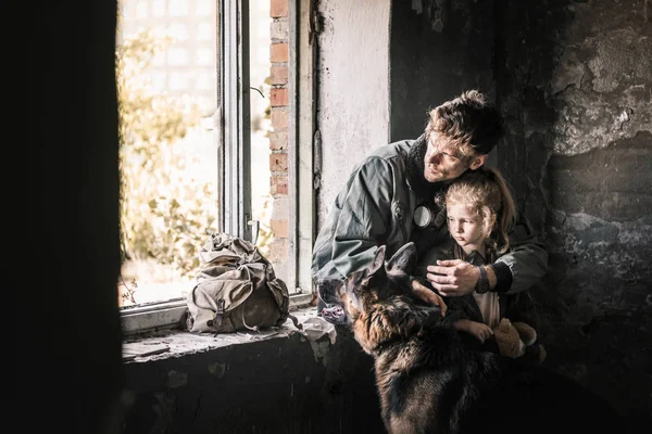 Мужчина обнимает ребенка возле немецкой овчарки в заброшенном здании, пост-апокалиптическая концепция — стоковое фото