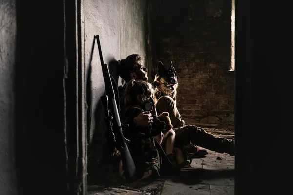 Мужчина и ребенок в противогазе сидят на полу рядом с пистолетом и немецкой овчаркой, пост-апокалиптическая концепция — стоковое фото