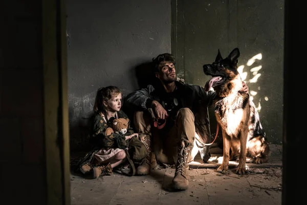 Handsome man and kid with teddy bear sitting on floor with german shepherd dog, post apocalyptic concept — Stock Photo