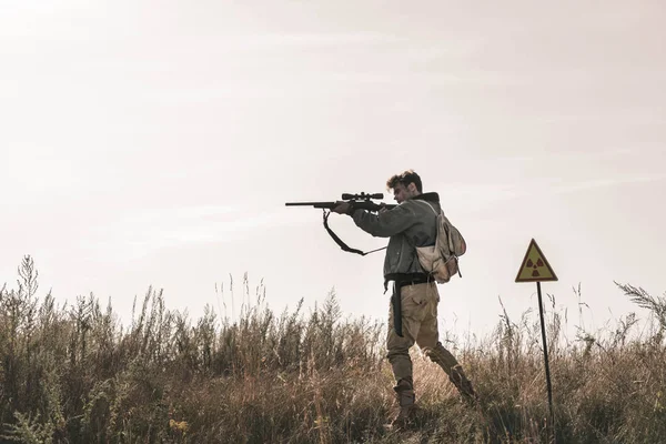 Schöner Mann mit Waffe in der Nähe toxischer Symbole im Feld, postapokalyptisches Konzept — Stockfoto