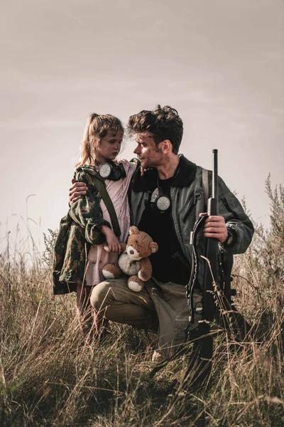 Bel homme tenant pistolet près de l'enfant avec ours en peluche sur le terrain, concept post-apocalyptique — Photo de stock