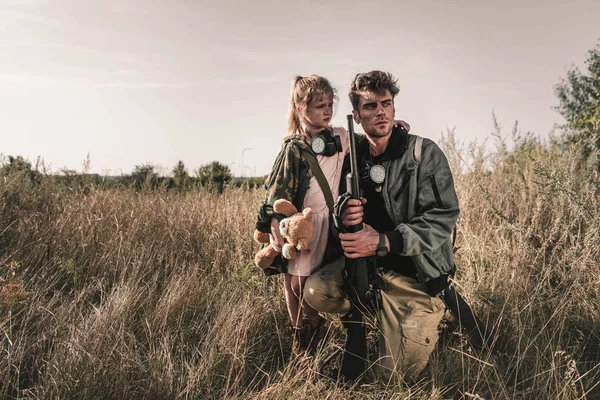 Schöner Mann mit Waffe in der Nähe von Kind mit Plüschtier im Feld, postapokalyptisches Konzept — Stockfoto