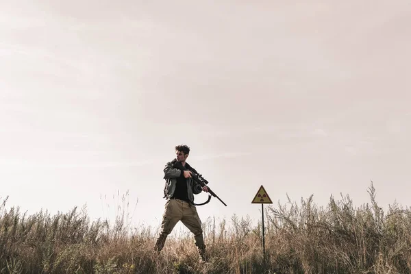 Hombre sosteniendo arma cerca de símbolo tóxico en el campo, concepto post apocalíptico - foto de stock