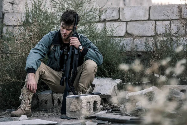 Foco seletivo de homem bonito sentado com arma perto da parede de tijolo fora, conceito pós-apocalíptico — Fotografia de Stock