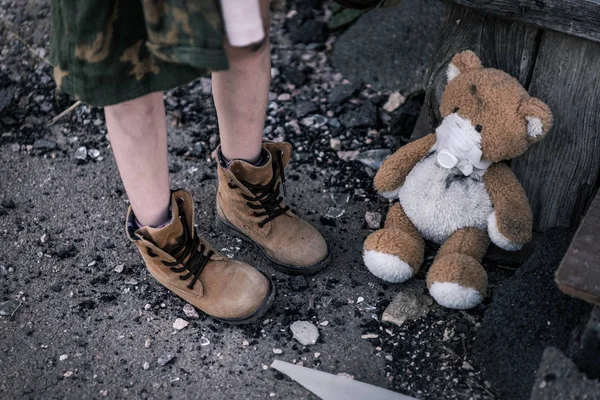 Cropped view of kid in boots standing near teddy bear on ground, post apocalyptic concept — Stock Photo