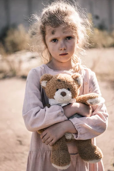 Niedliches Kind mit schmutzigem Gesicht, das in die Kamera schaut, während es Teddybär hält, postapokalyptisches Konzept — Stockfoto