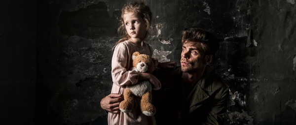 Panoramic shot of upset man near kid with teddy bear in dirty room, post apocalyptic concept — Stock Photo