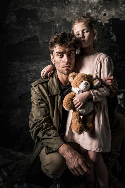 Handsome man sitting near kid with teddy bear in dirty room, post apocalyptic concept — Stock Photo