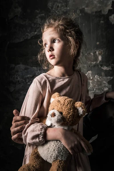 Обрезанный вид человека, трогающего грустного ребенка с плюшевым мишкой в грязной комнате, постапокалиптическая концепция — стоковое фото