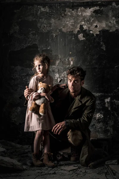 Handsome man sitting near upset kid with teddy bear in dirty room, post apocalyptic concept — Stock Photo