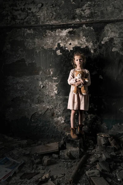 Triste niño de pie y sosteniendo osito de peluche en la habitación sucia, concepto post apocalíptico - foto de stock