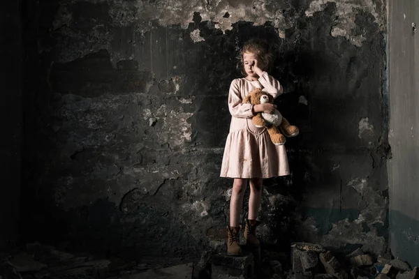 Frustrado niño llorando mientras sostiene oso de peluche en la habitación sucia, concepto post apocalíptico - foto de stock