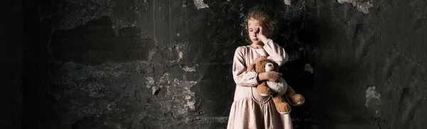 Panoramic shot of frustrated kid crying while holding teddy bear in dirty room, post apocalyptic concept — Stock Photo