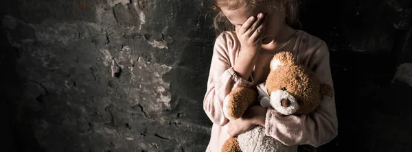 Panoramic shot of upset kid touching face while holding teddy bear in dirty room, post apocalyptic concept — Stock Photo