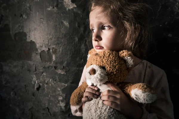 Aufgebrachtes Kind hält Teddybär in schmutzigem Raum, postapokalyptisches Konzept — Stockfoto