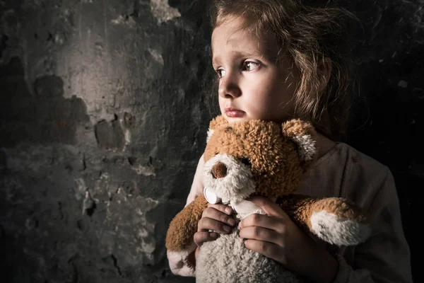 Niño molesto sosteniendo osito de peluche en la habitación sucia, concepto post apocalíptico - foto de stock