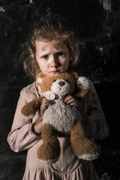 Frustrado niño sosteniendo osito de peluche en habitación sucia, concepto post apocalíptico - foto de stock