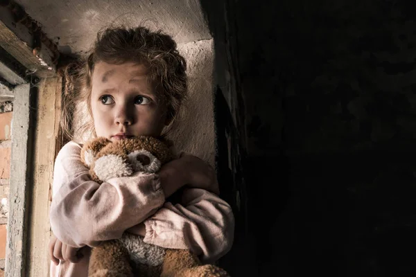 Расстроенный ребенок держит плюшевого мишку в грязной комнате, пост-апокалиптическая концепция — стоковое фото